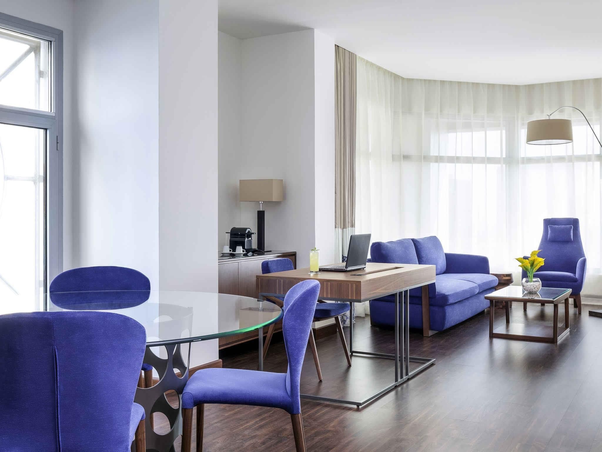 A modern living room with a glass coffee table, blue sofas, and a dining area with a small kitchen setup.