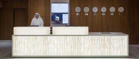 A reception desk with a digital screen displaying room information, a man standing behind it, and five clocks on the wall.