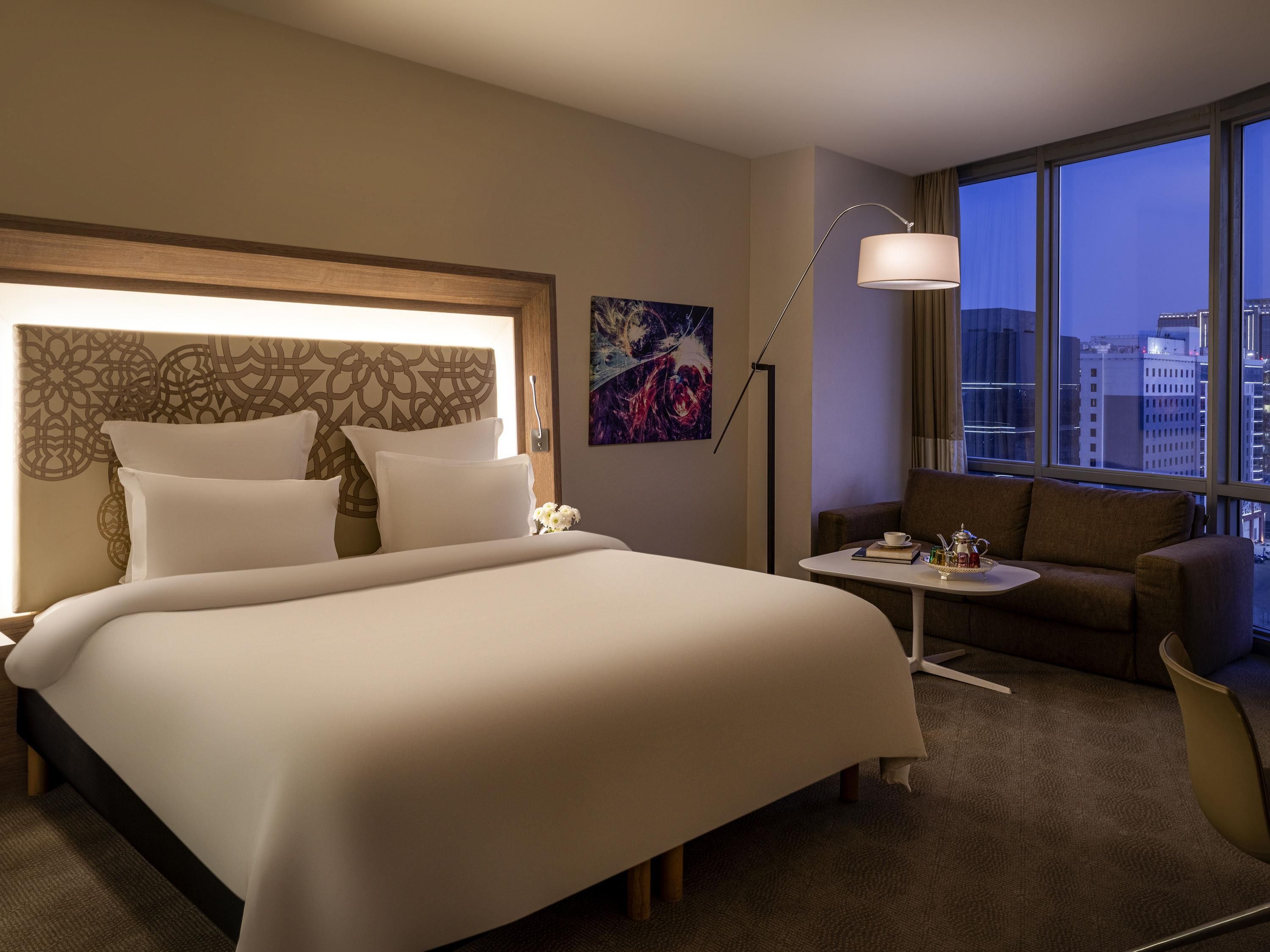 A hotel room with a large bed, a small table with a cup and book, a couch, a floor lamp, and a city view through large windows.