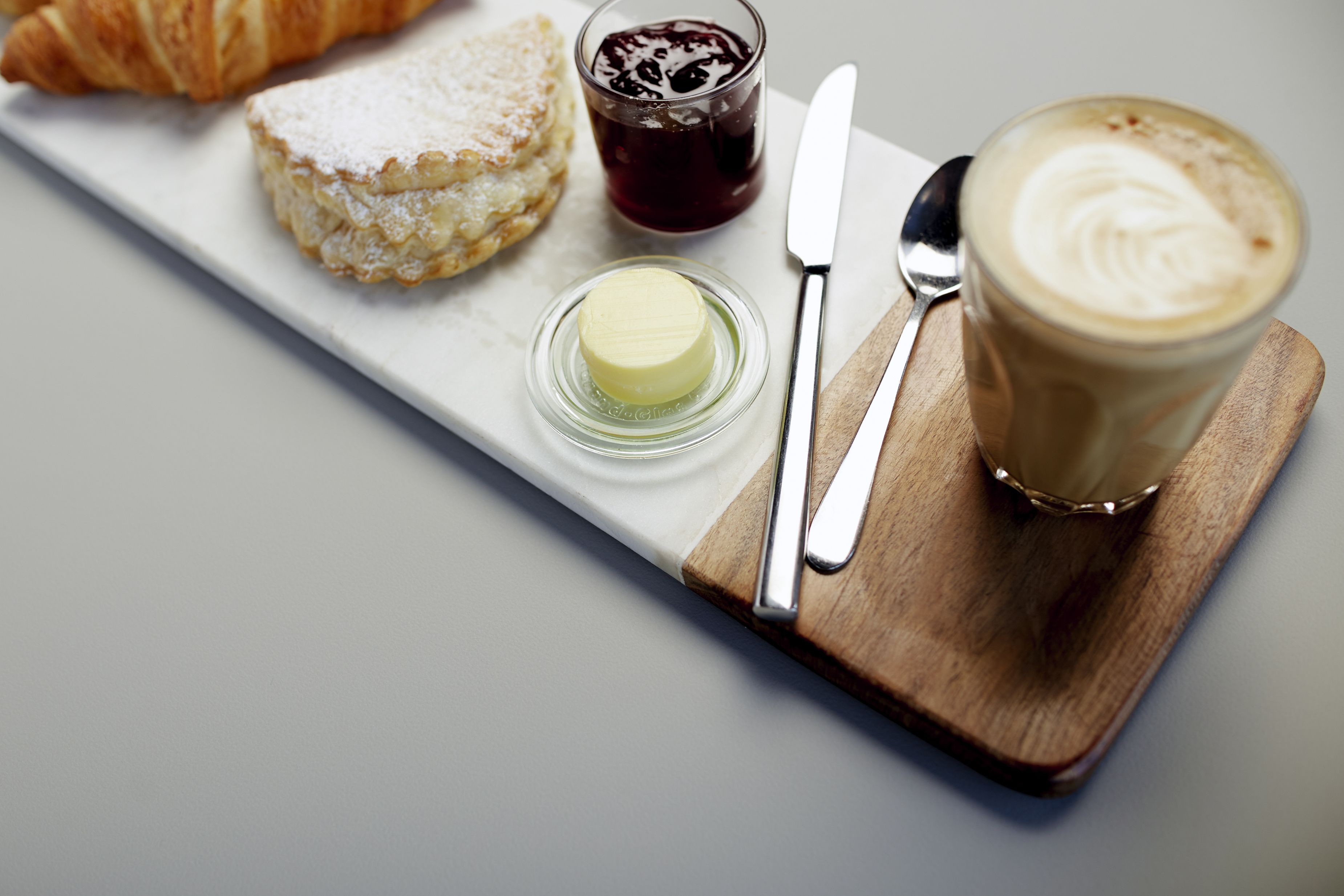 Een ontbijtplateau met een croissant, boter, jam en een glas koffie op een houten plank.