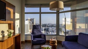 A modern living room with a city view, a wooden desk, a blue armchair, a round coffee table with a bowl of apples, and a hanging lamp. - Novotel Jeddah Tahlia (Jeddah)