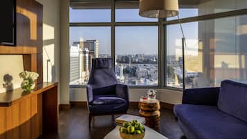 A modern living room with a city view, a wooden desk, a blue armchair, a round coffee table with a bowl of apples, and a hanging lamp.