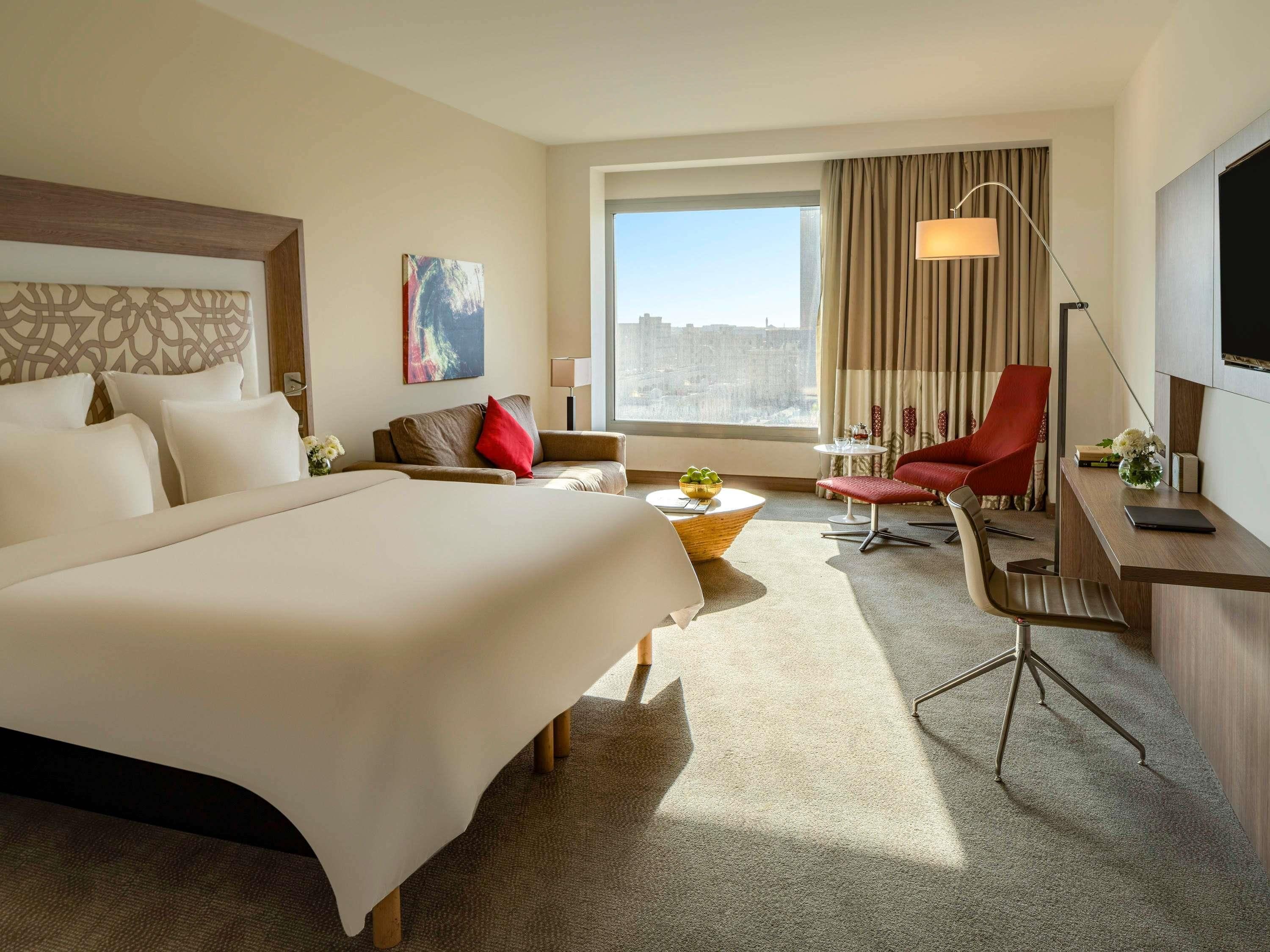 Una habitación de hotel con una cama grande, un escritorio con una silla, una silla roja, una mesita con un cuenco, una lámpara y un cuadro en la pared.