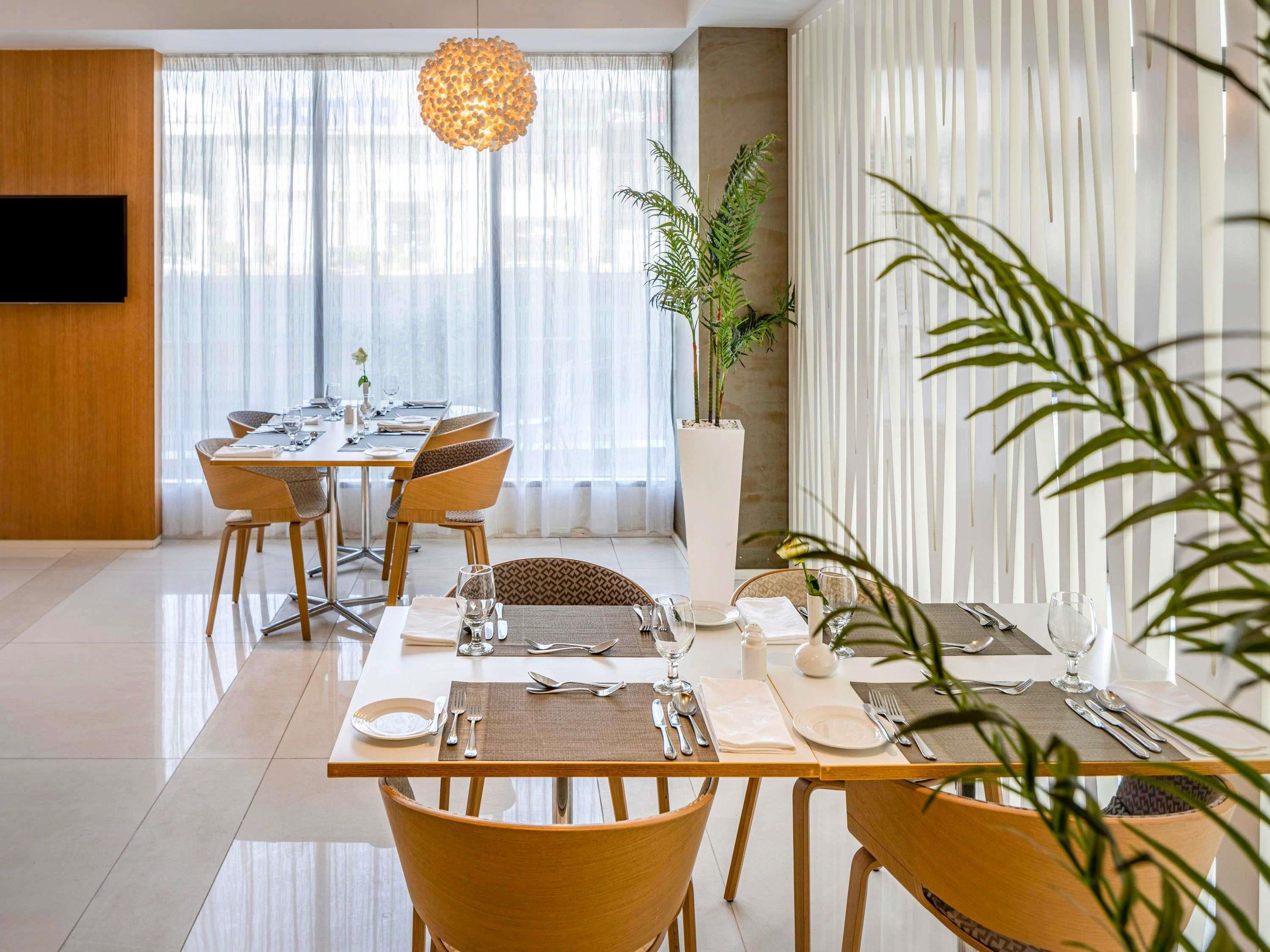 A dining area with a long table, chairs, a potted plant, and a hanging light fixture.
