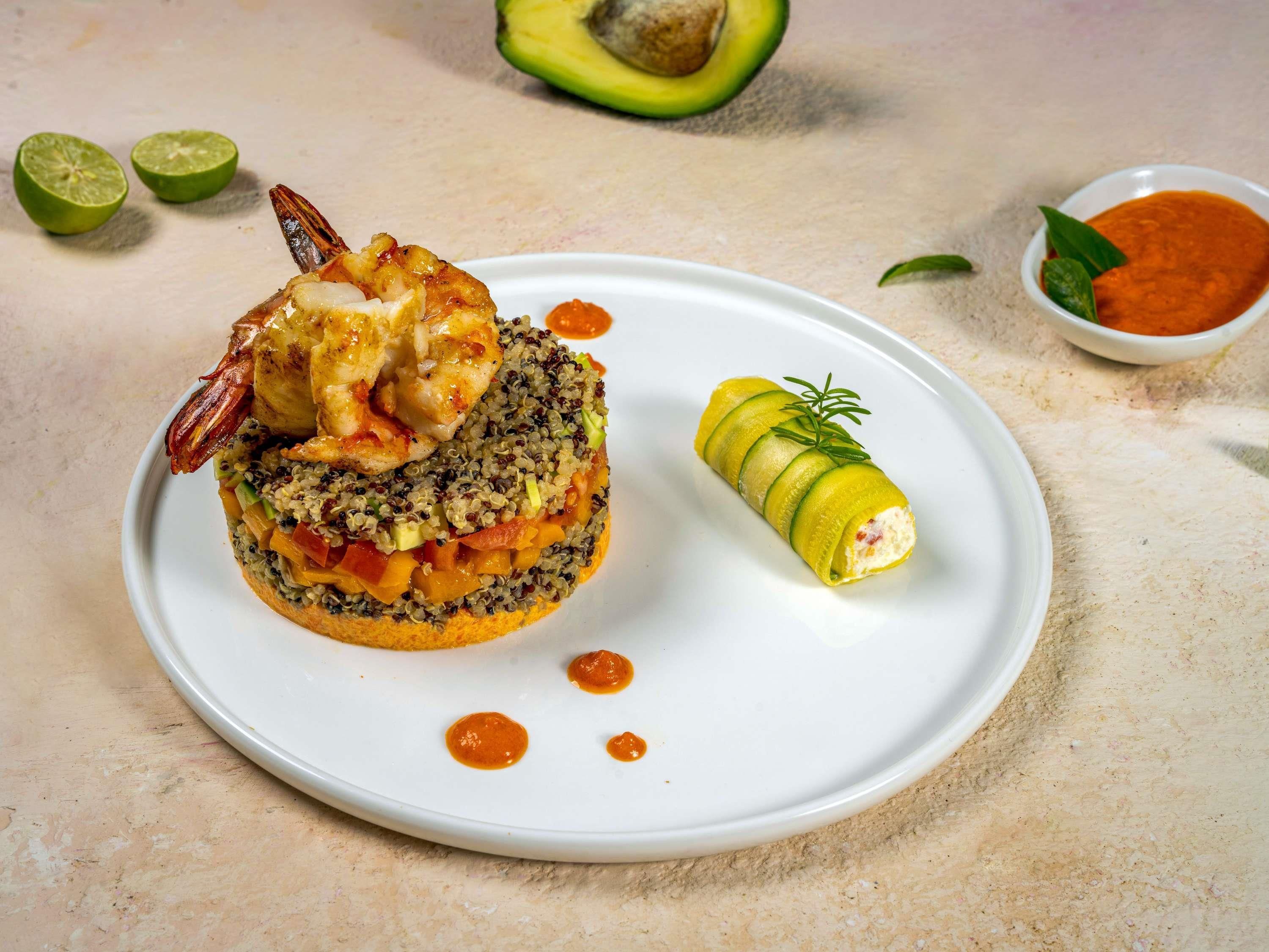 A plate with a quinoa-based dish, a rolled vegetable, and a small bowl of sauce.
