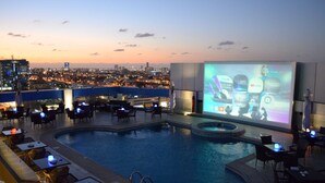 A rooftop pool area with a cityscape view at dusk. There is a projection screen displaying various apps and services. - Novotel Jeddah Tahlia (Jeddah)