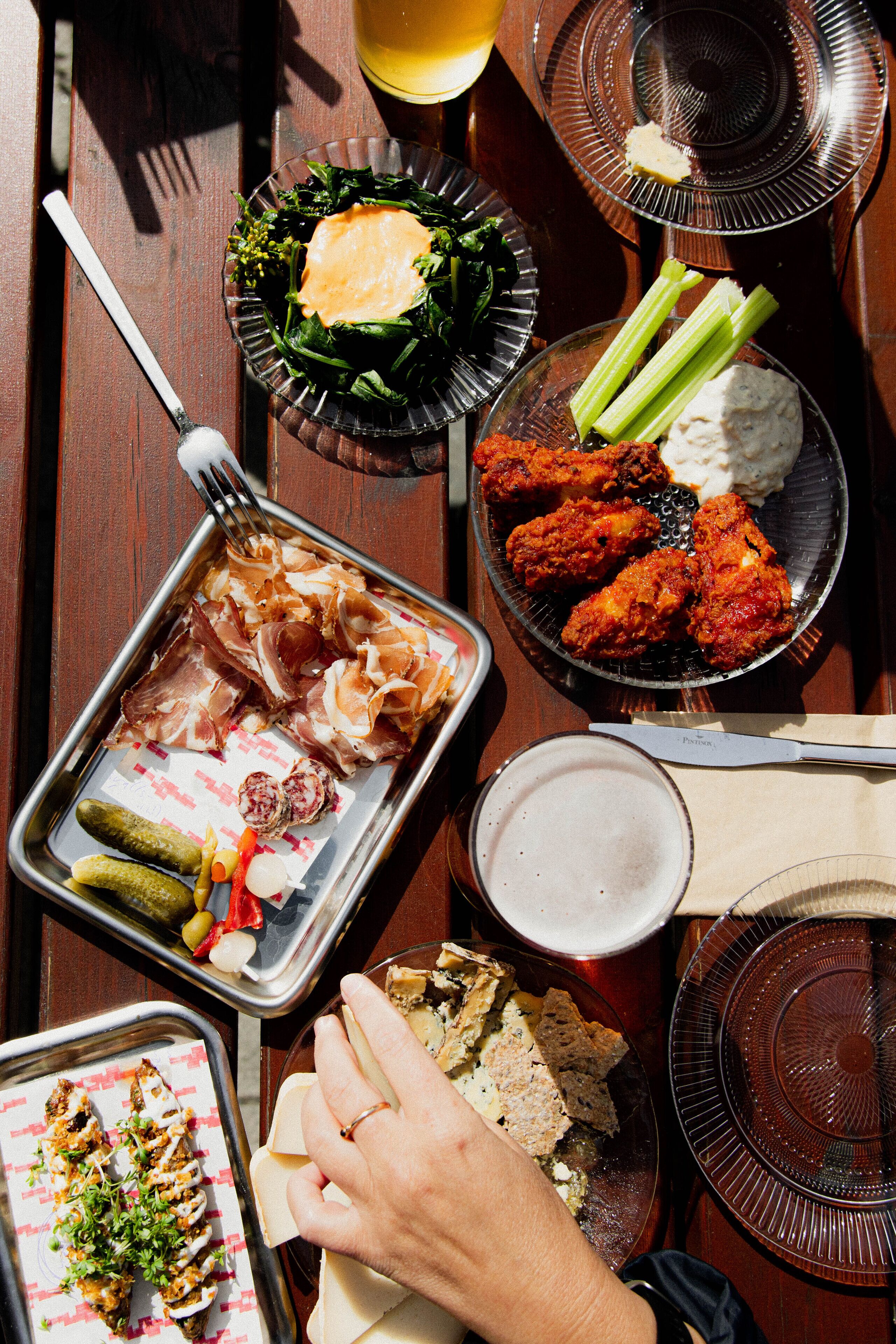 Variedad de alimentos sobre una mesa de madera, incluyendo una bandeja con carnes, un bol con alitas de pollo y un plato con pepinillos y verduras.
