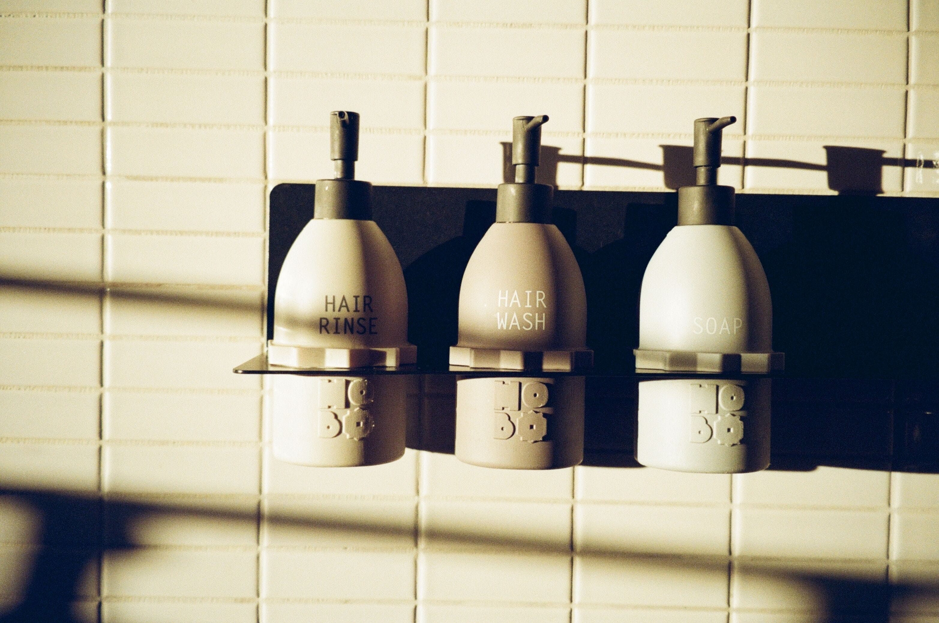 Een badkamer met drie flessen, gelabeld 'HAIR RINSE', 'HAIR WASH' en 'SOAP', bevestigd aan een betegelde muur.