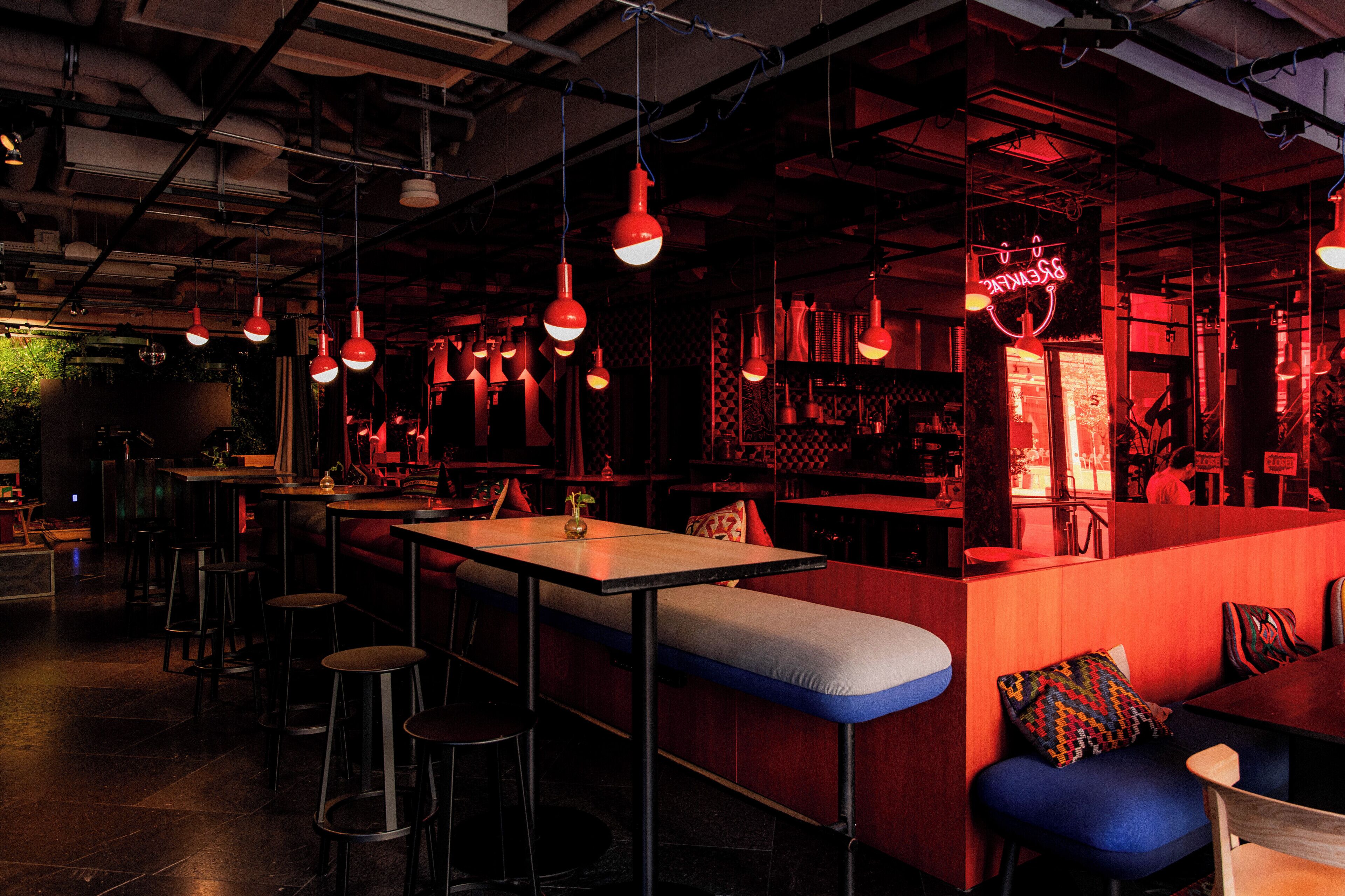 A dimly lit restaurant with red and blue seating, hanging red pendant lights, and a neon sign.