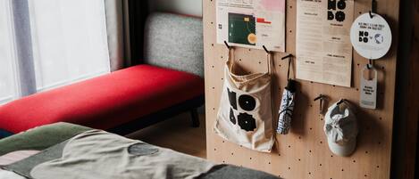 Una habitación ordenada con una cama, un banco rojo y un panel de pegboard con varios objetos como un mapa, una bolsa y un sombrero.