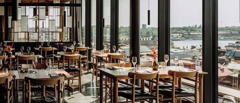 A restaurant with large windows offering a view of a river and buildings. Tables are set with wine glasses and cutlery.