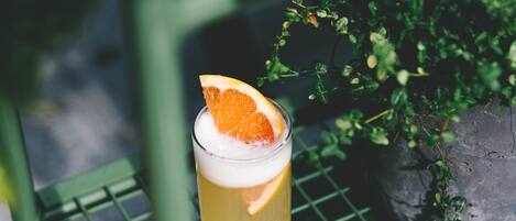 A glass of yellow beverage with an orange slice garnish on a metal shelf, surrounded by greenery.