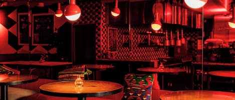 Un bar tenuemente iluminado con mesas redondas y asientos rojos.