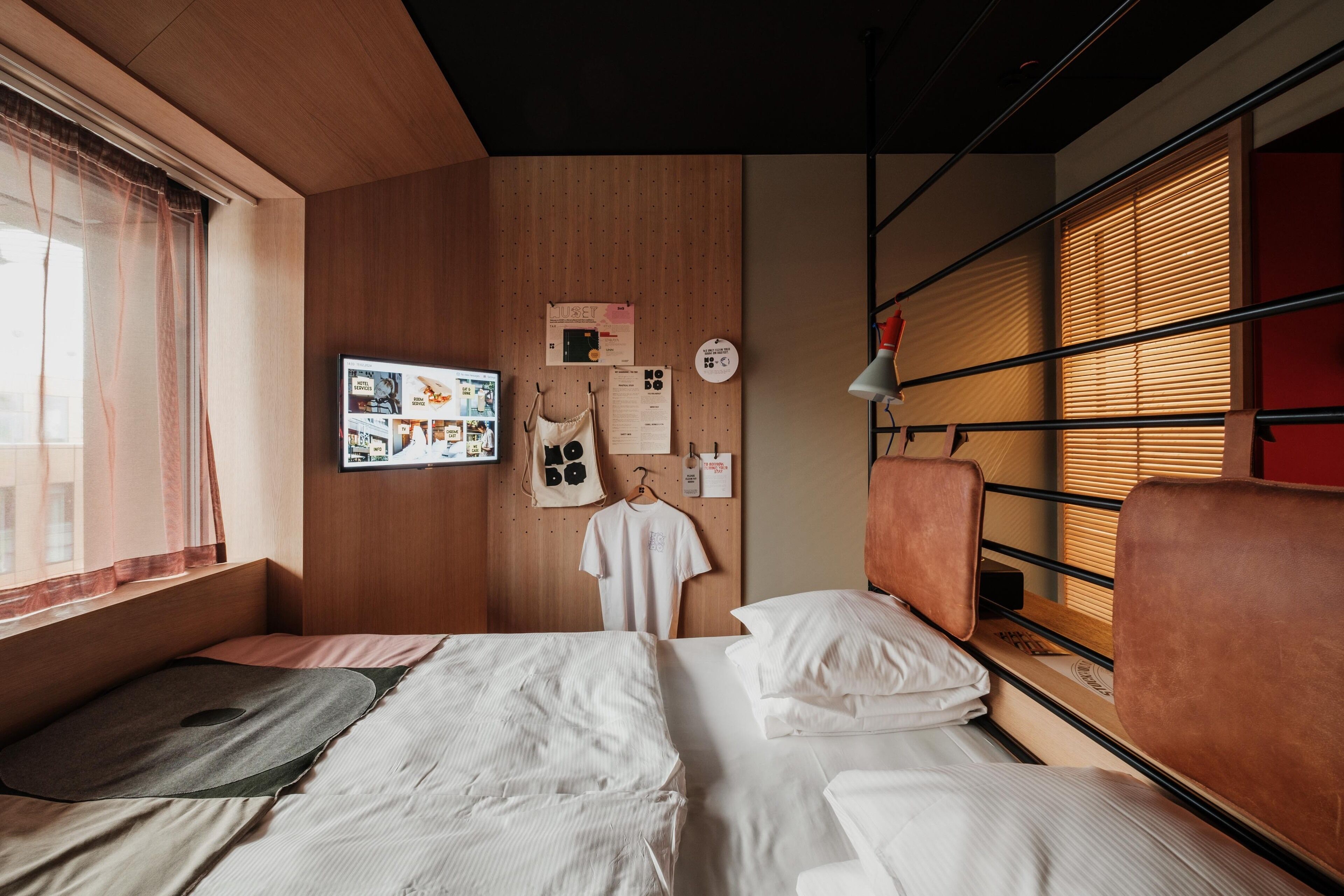 A hotel room with two beds, a wooden wall, a TV, and a window with blinds.