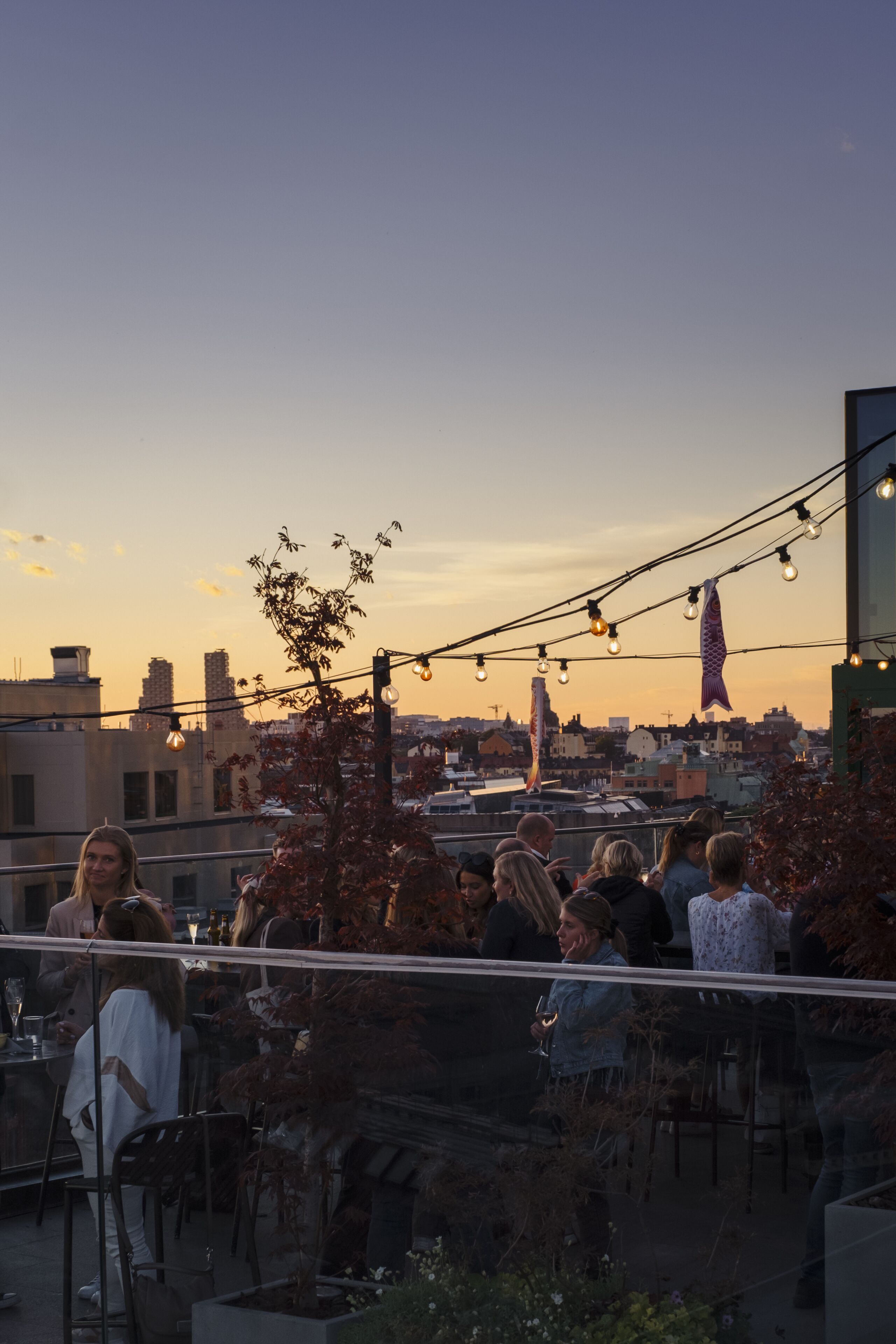 Una terrazza sul tetto con tavoli e sedie, circondata da edifici cittadini, illuminata da luci decorative.