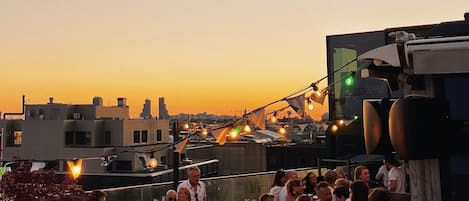 A rooftop bar with tables and chairs, overlooking a cityscape at sunset.