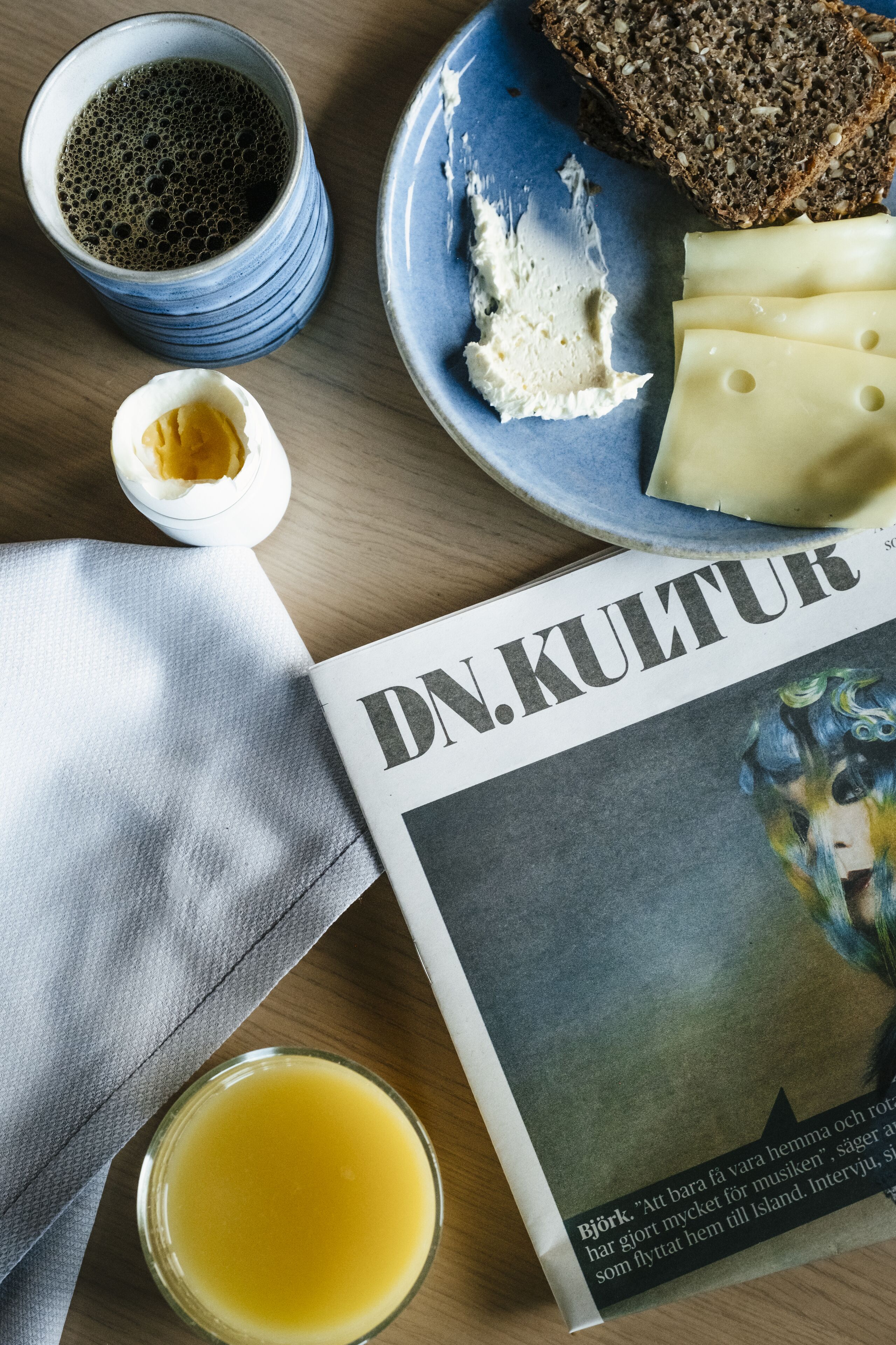 A table with a plate of cheese, a slice of bread, a cup of coffee, a glass of juice, and a magazine titled 'DN. KULTUR'.