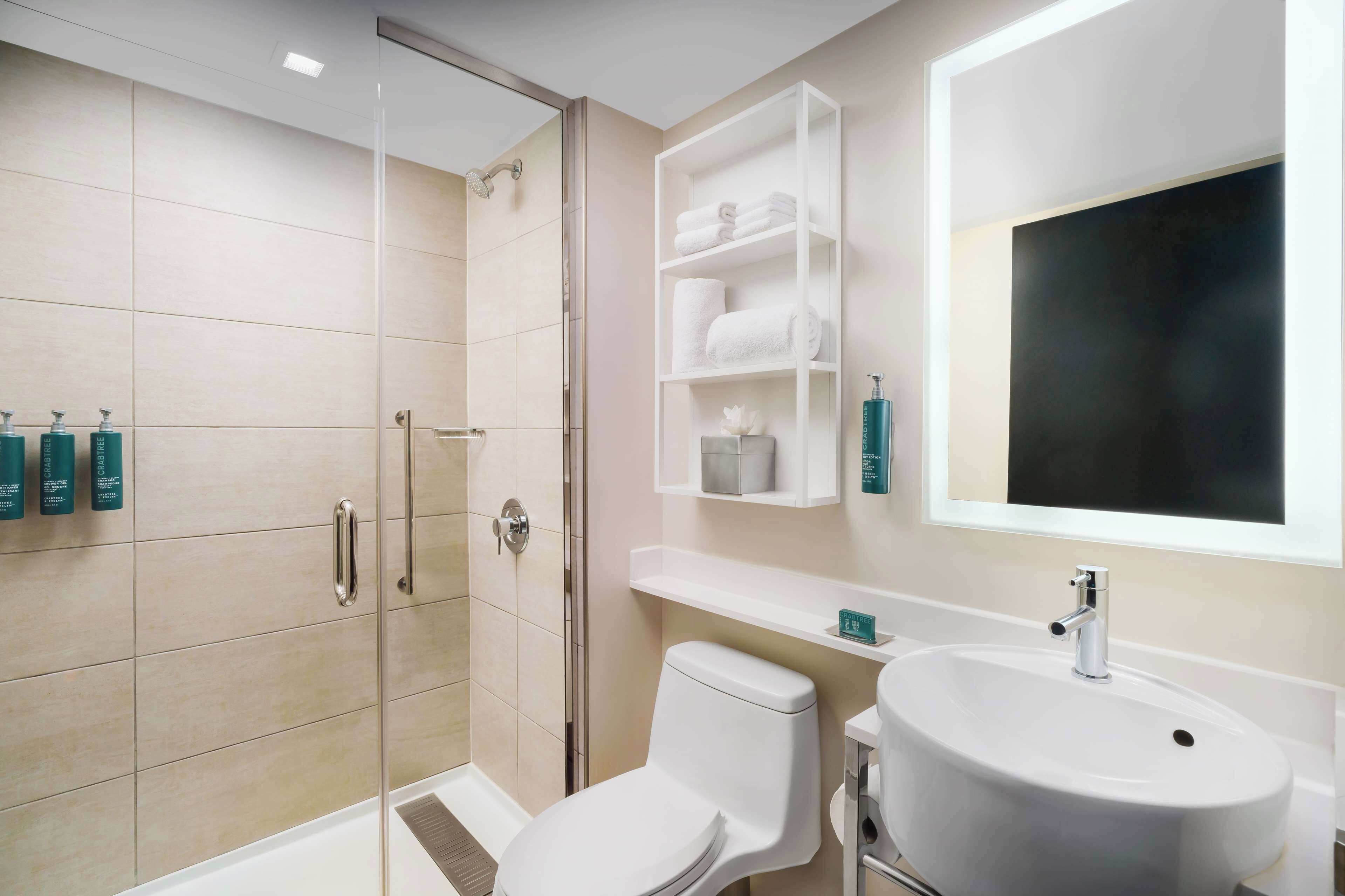 A modern bathroom with a glass shower enclosure, a white toilet, and a sink with a mirror above it. There are shelves with towels and toiletries.