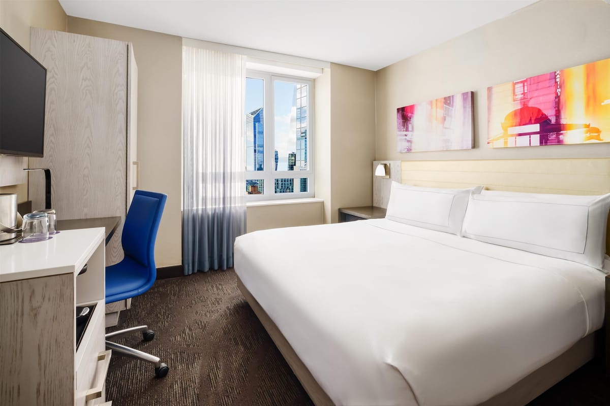 Quarto de hotel com uma cama grande, uma mesa com cadeira, uma televisão e uma janela com cortinas.