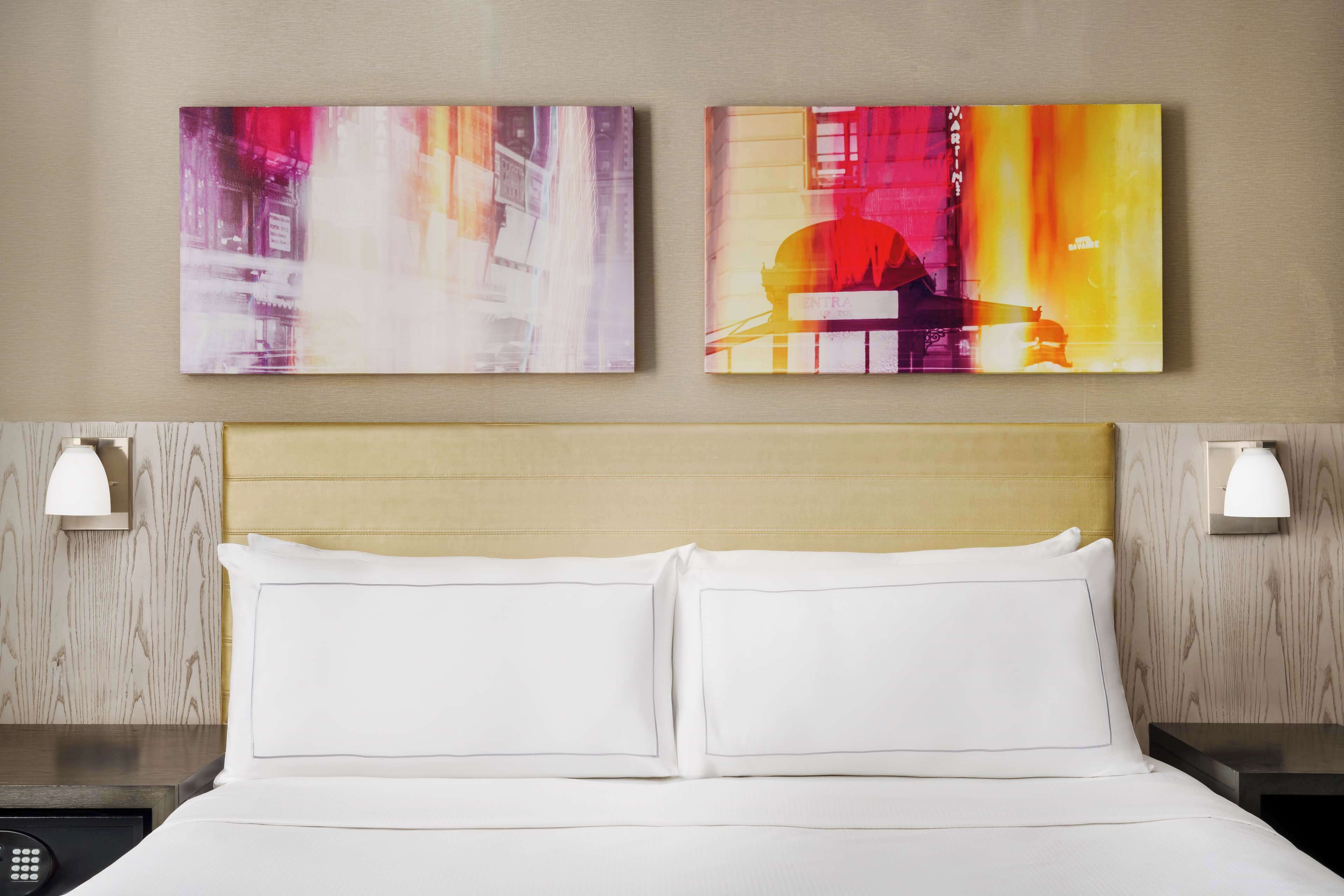 A hotel room with a bed, two wall-mounted lamps, and two abstract paintings on the wall.