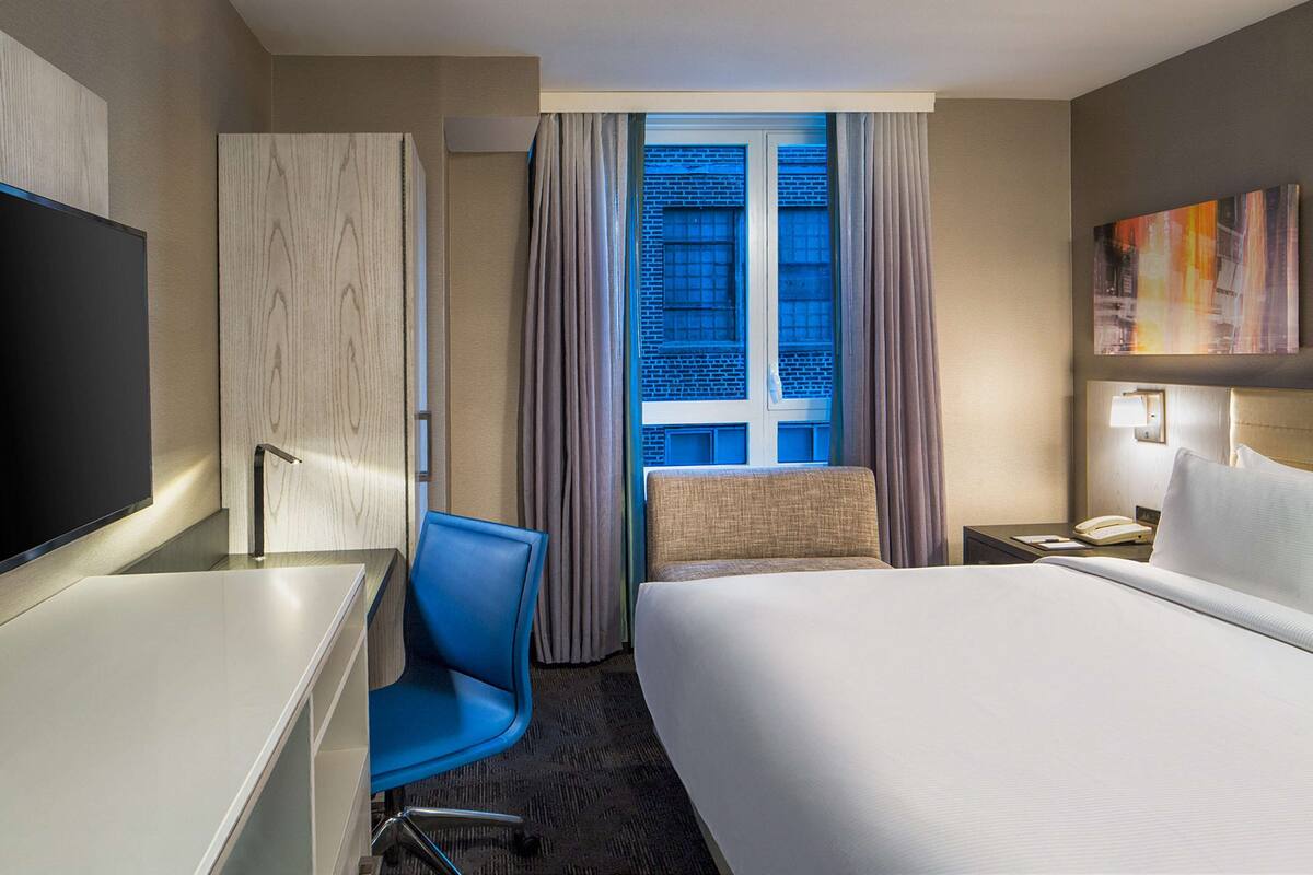 Quarto de hotel com cama, escrivaninha, cadeira, televisão, janela com cortinas e um quadro na parede.