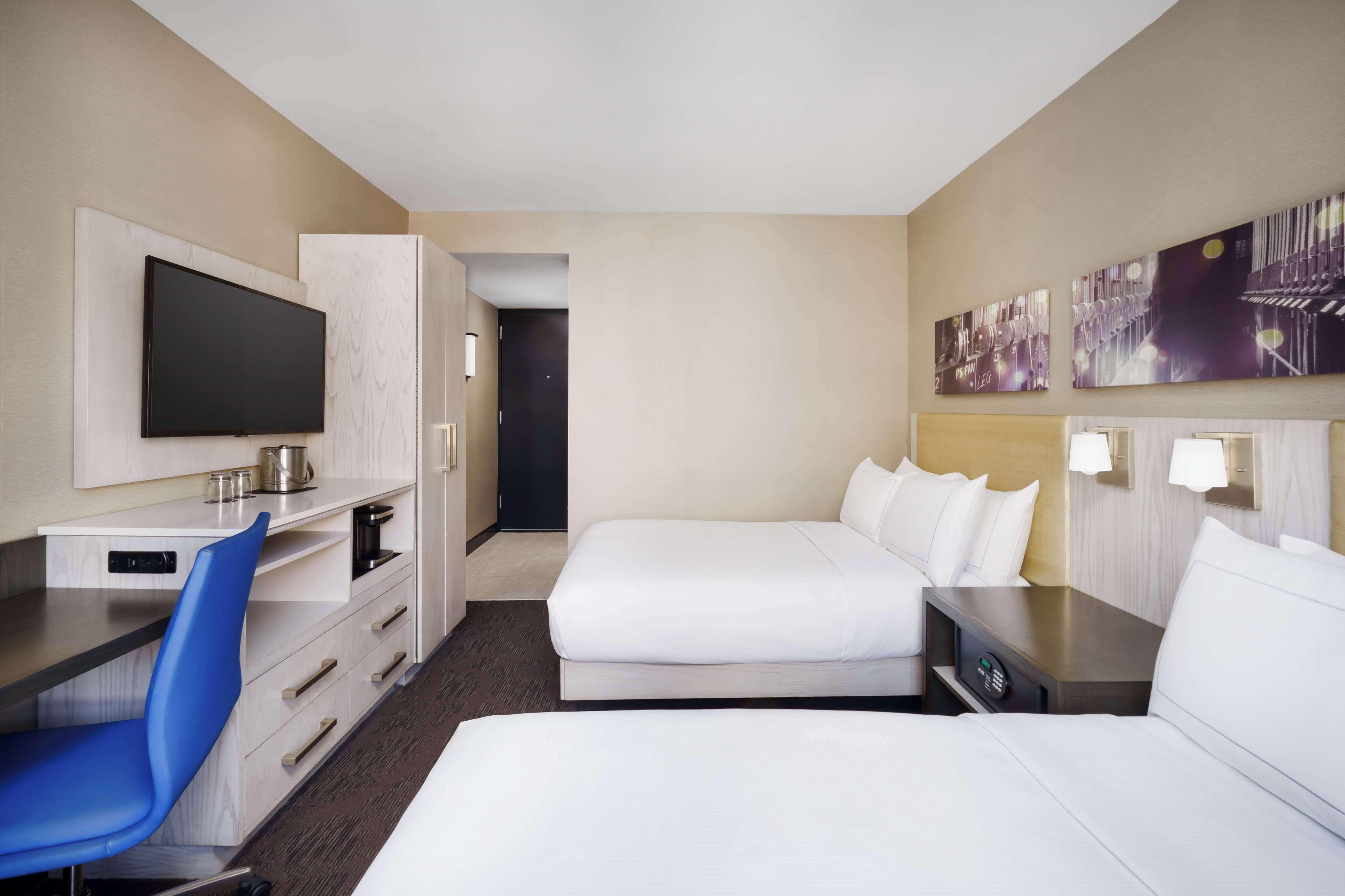 A hotel room with a flat-screen TV, a desk with a chair, two beds, and artwork on the wall.