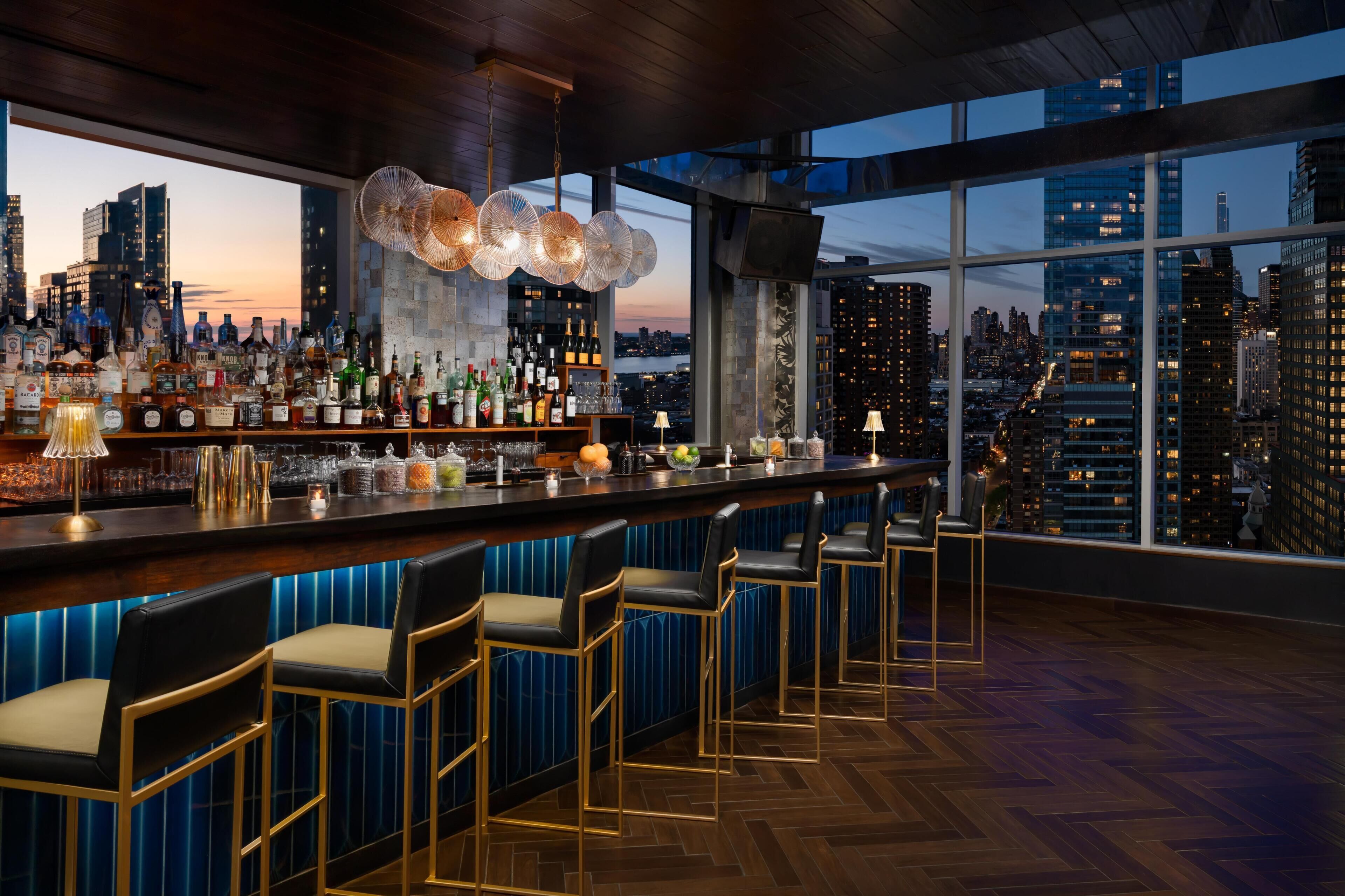 A bar with a city view, bar stools, and a variety of bottles.