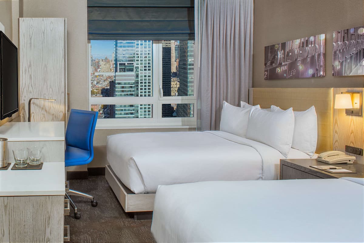 Quarto de hotel com duas camas, uma mesa, uma cadeira, televisão, janela com vista para a cidade e obras de arte na parede.