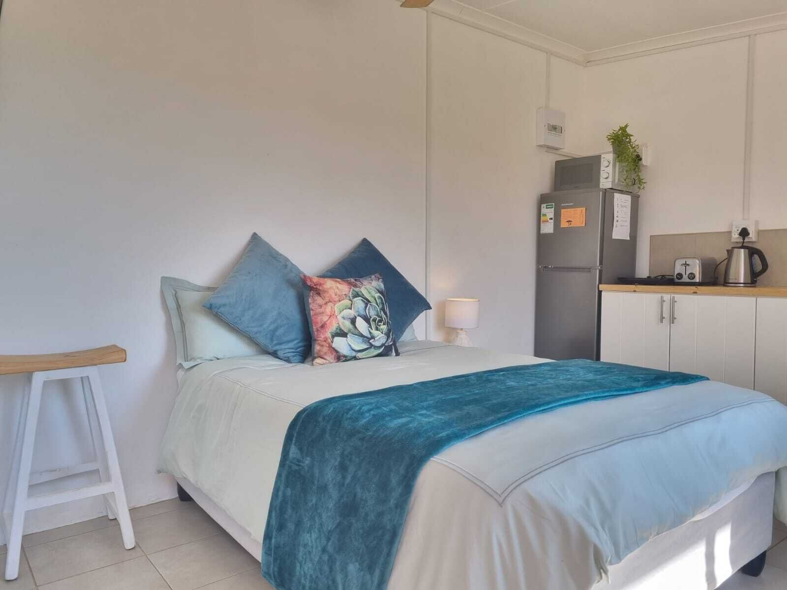 A bedroom with a bed, pillows, a bedside table, a nightstand, a small kitchenette with a fridge, a kettle, and a toaster.
