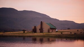 A house with a green roof and a chimney, situated by a body of water with mountains in the background. - Kwetu Guest Farm (Swellendam)