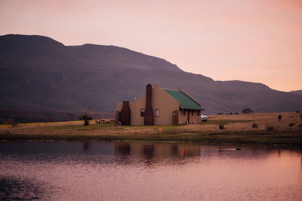 A house with a green roof and a chimney, situated by a body of water with mountains in the background.