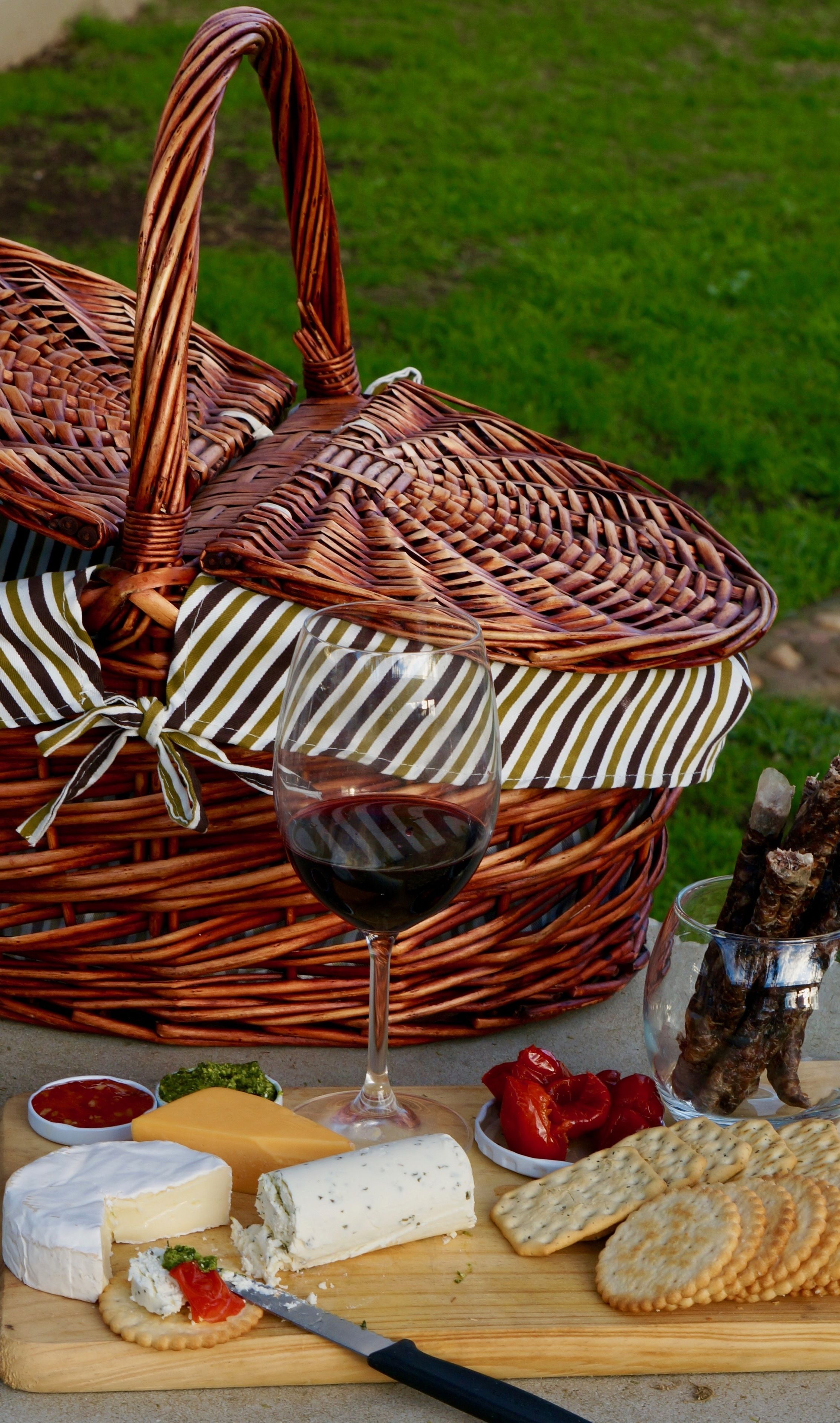 A wicker picnic basket with a wine glass, cheese, crackers, and charcuterie on a wooden board.