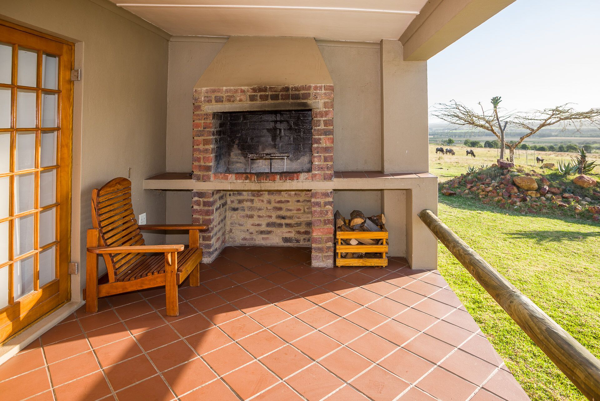 Un patio con una chimenea de ladrillo, una silla de madera y vistas a un campo cubierto de césped con animales.