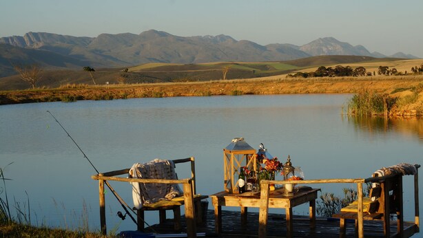A wooden dock with chairs and a table by a calm lake, surrounded by mountains. - Kwetu Guest Farm (Swellendam)