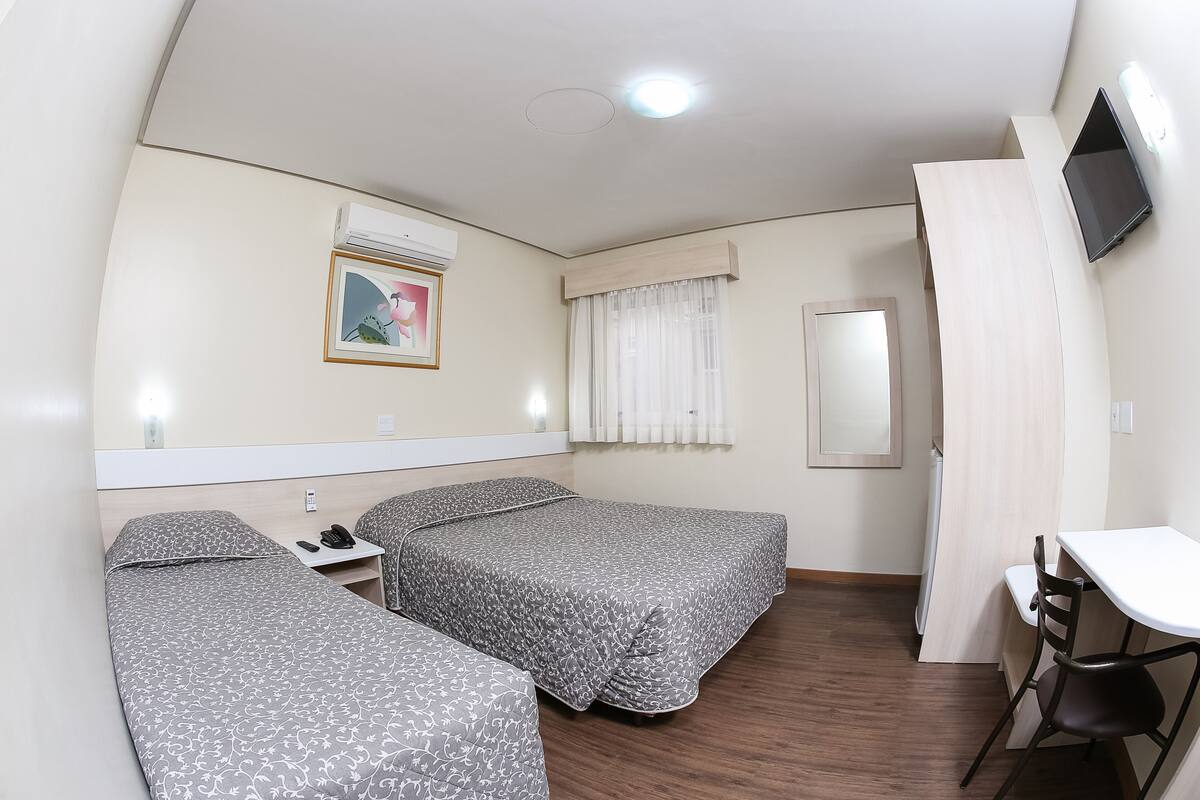 A hotel room with two single beds, a desk, a chair, a TV mounted on the wall, and a window with curtains.