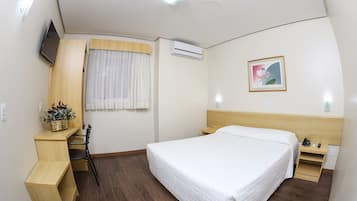 A hotel room with a bed, a desk, a chair, a small table, a nightstand, a wall-mounted TV, an air conditioner, a window with curtains, a picture on the wall, and a plant on the desk.