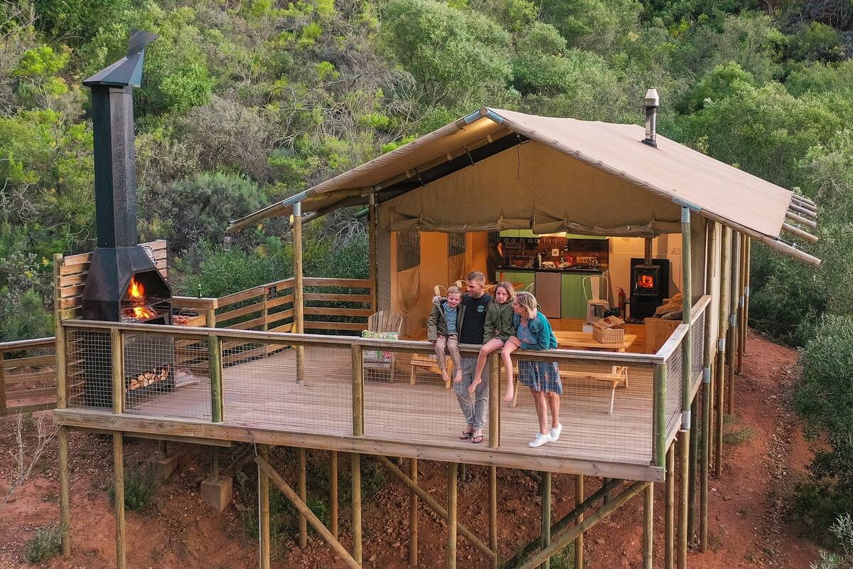 Eine Holzterrasse mit überdachter Sitzecke, Kamin und Blick auf einen bewaldeten Hügel.