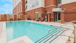 Piscina com deck ladrilhado, cadeiras de descanso e um prédio de tijolos ao fundo.