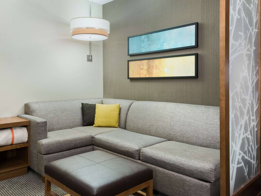 A modern hotel room with a grey sectional sofa, a wooden side table with a towel, a bench, and two abstract paintings on the wall.