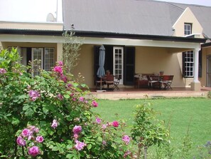 A house with a covered porch, a lawn, and blooming flowers. - Die Waenhuis Guest House (George)