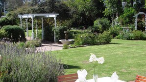 A garden with a white pergola, a fountain, and a table set for dining with white tablecloths and napkins. - Die Waenhuis Guest House (George)