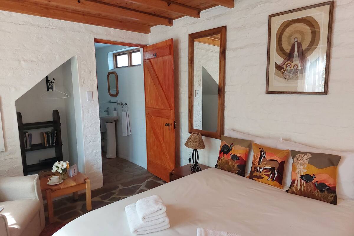 Een slaapkamer met een bed, kussens, een nachtkastje, een lamp, een boekenkast, en een badkamer die door een open deur zichtbaar is.