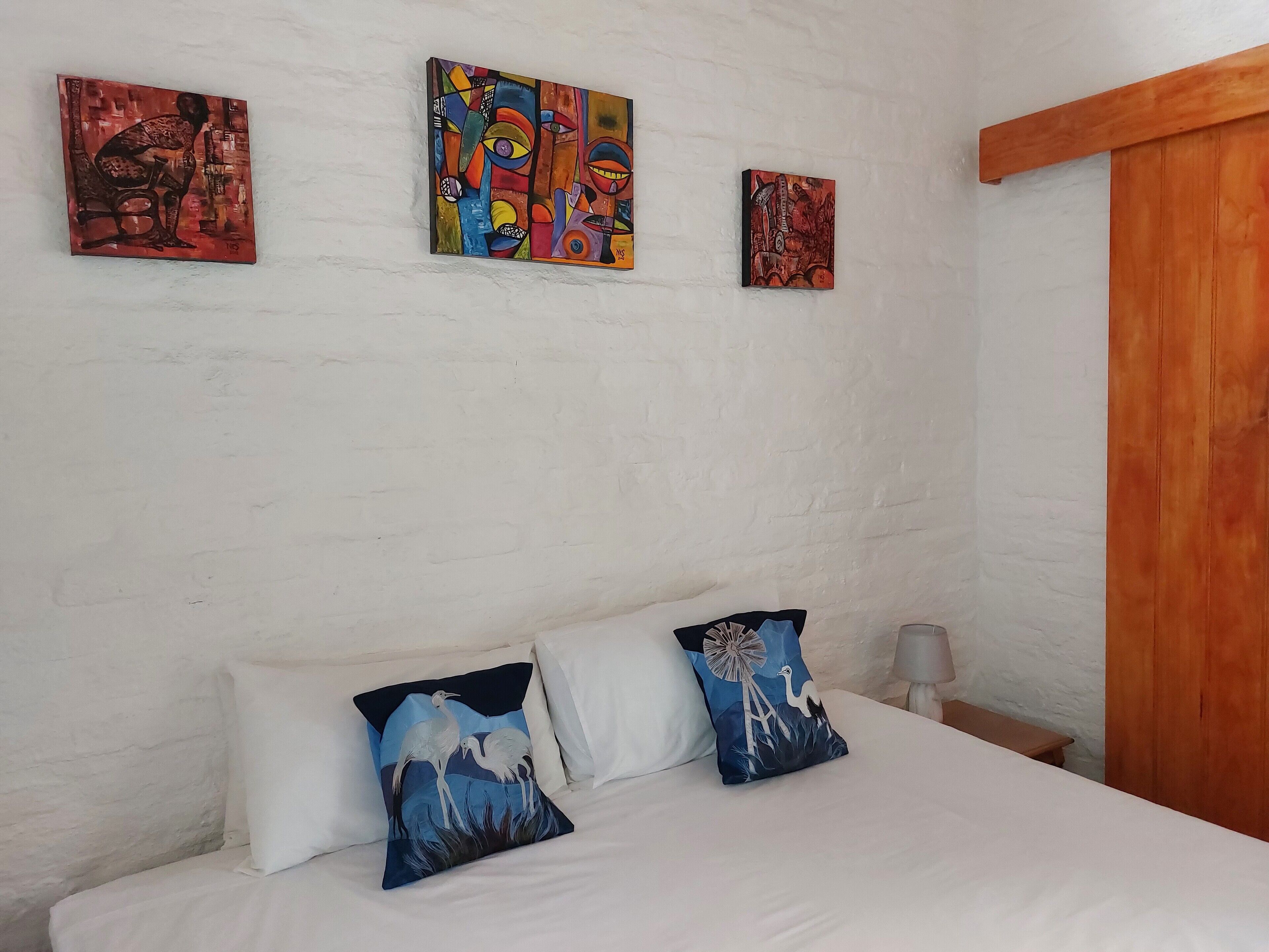 A bedroom with a bed, white pillows, and three colorful paintings on the wall.
