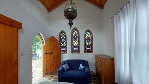 A room with a stone floor, a wooden ceiling, a blue sofa, a wooden dresser, and stained glass windows.