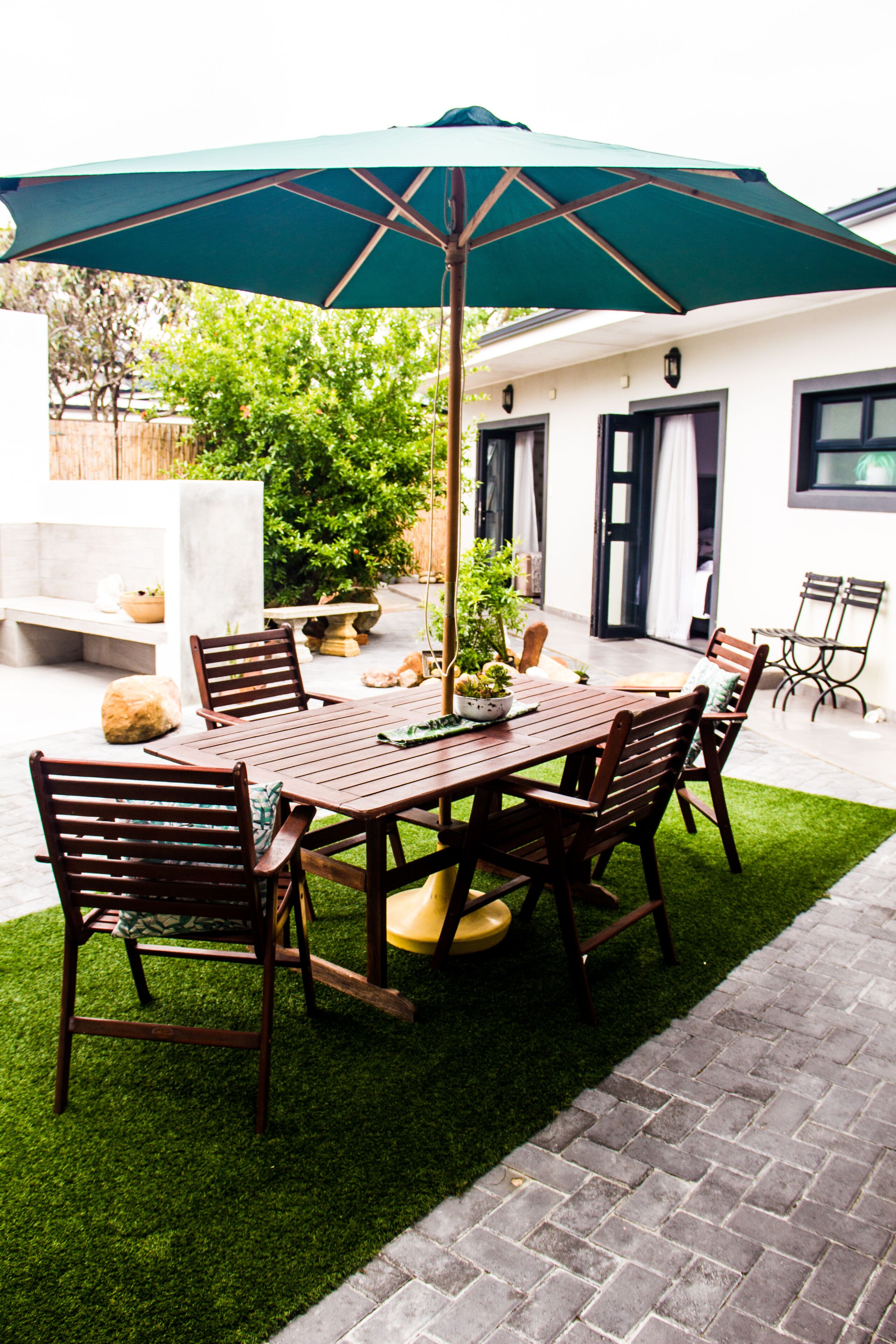 A patio with a wooden table and chairs, a green umbrella, and a small garden area with potted plants.