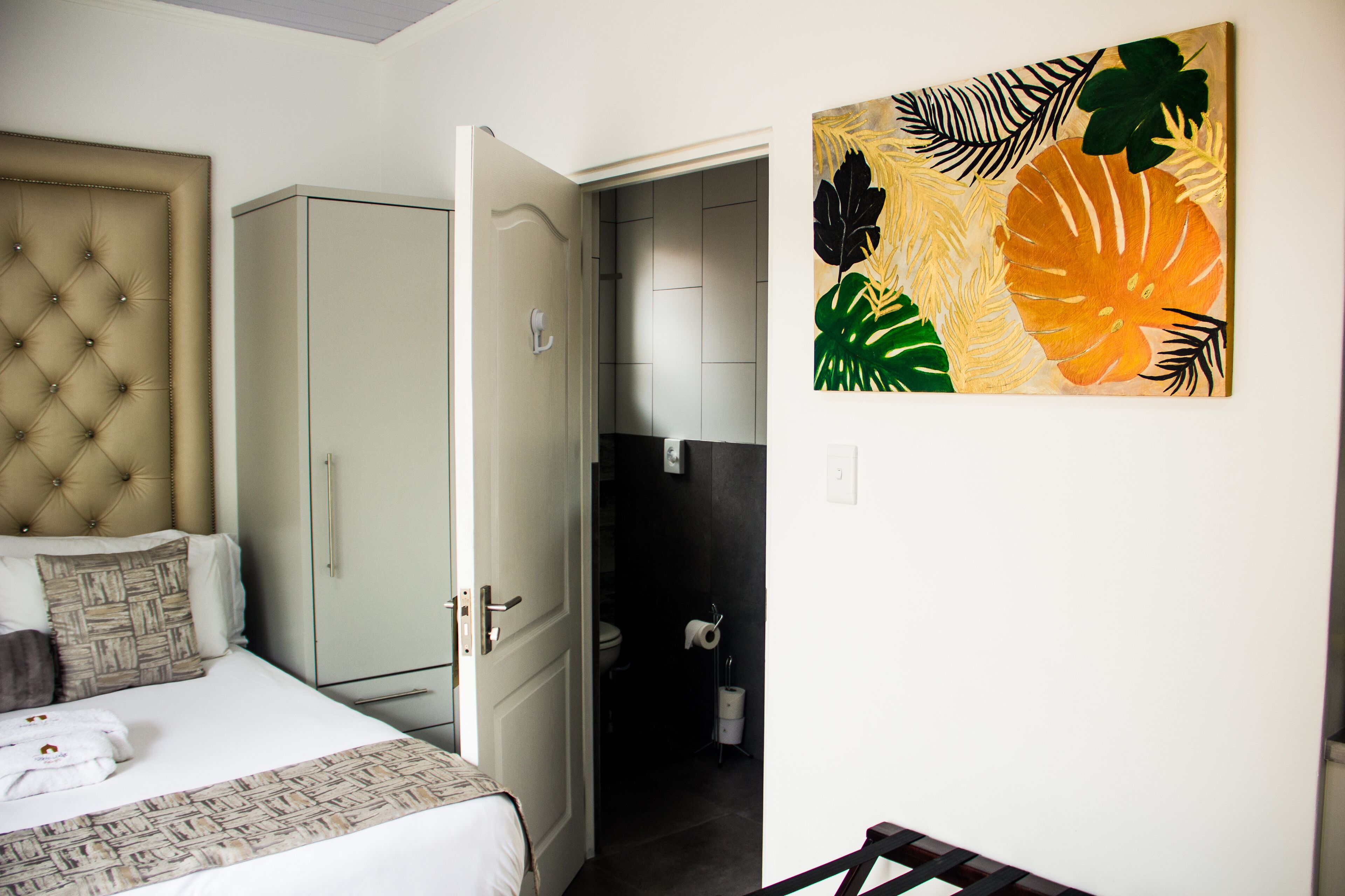 A bedroom with a bed, a wardrobe, a door leading to a bathroom, and a wall with a tropical leaf painting.