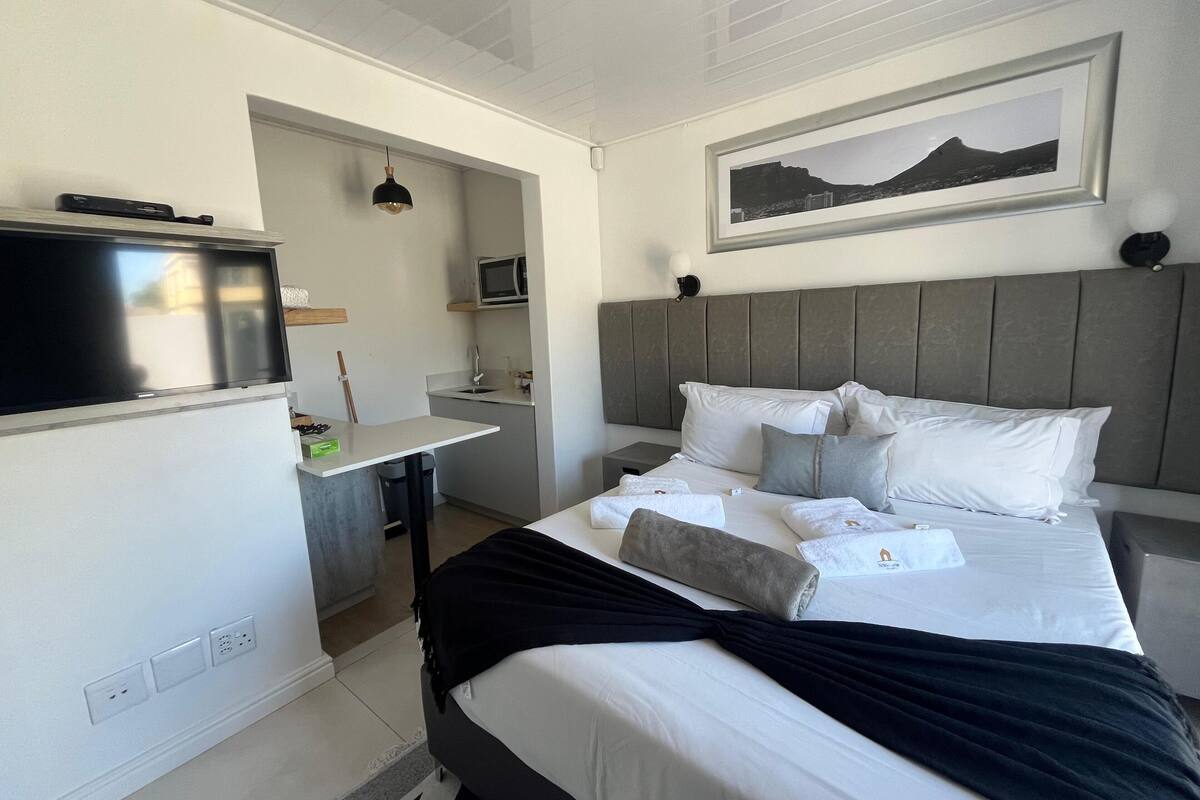 A modern hotel room with a bed, a flat-screen TV, a small kitchenette, and a mounted picture of a mountain landscape.