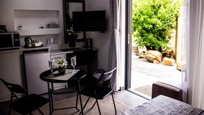 Habitación de hotel compacta con kitchenette, comedor con mesa y sillas, televisor de pantalla plana montado en la pared y puerta corrediza de vidrio que da a un área exterior con vegetación.