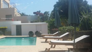 En swimmingpool med solstolar och parasoller, omgiven av en trädgård och en byggnad.