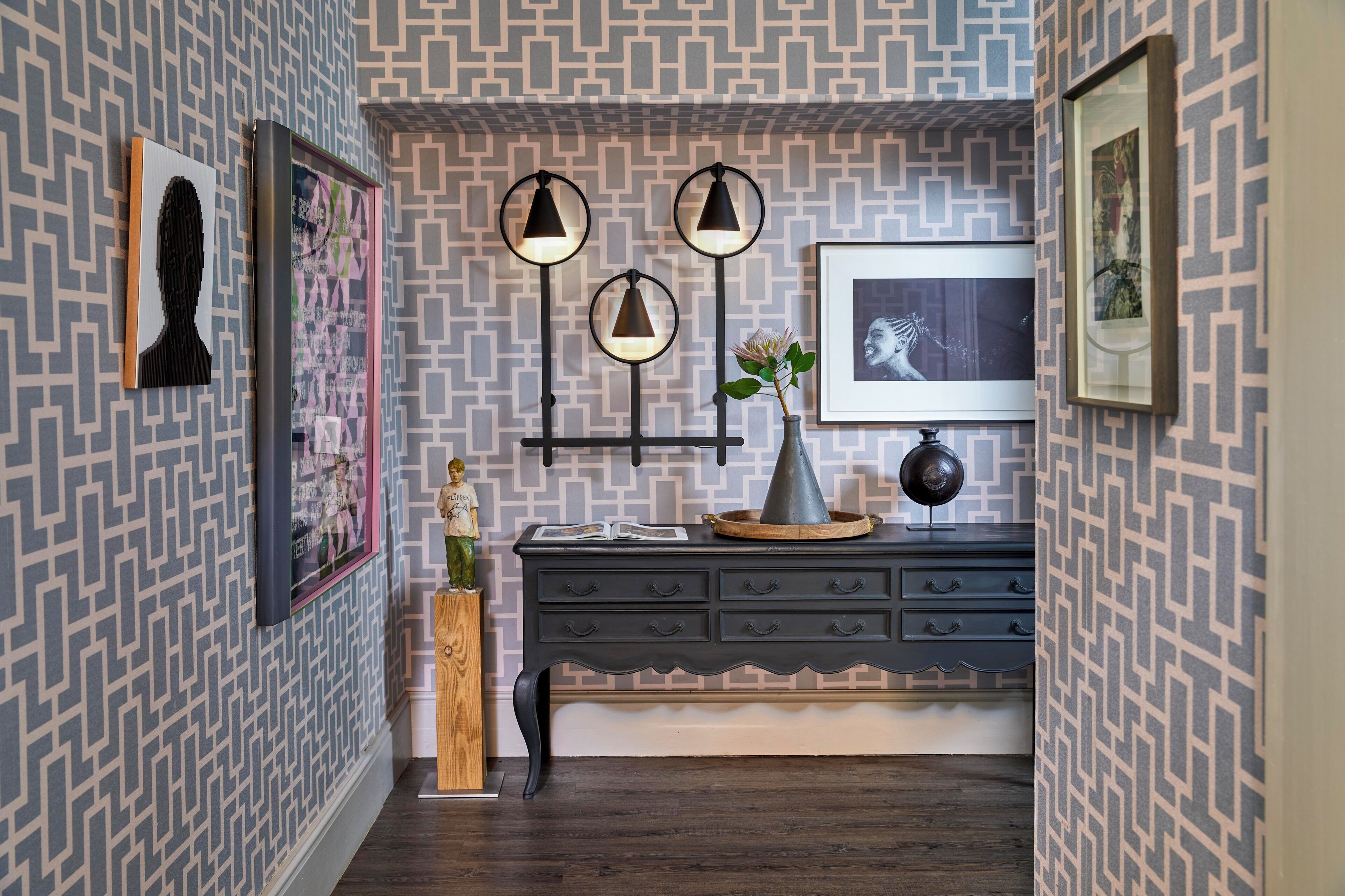A hallway with patterned wallpaper, a dark wooden floor, a black dresser with drawers, and wall-mounted lights.